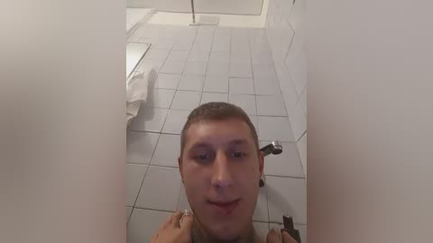 A video shows a young man with short brown hair and fair skin, smiling while taking a selfie in a white-tiled bathroom, with a white towel on the left and a metal shower fixture in the background.