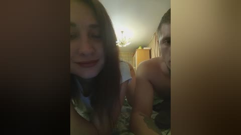 Video of a young woman with straight, brown hair, smiling, and a shirtless man in a dimly-lit bedroom with wooden furniture.