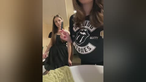 Video of a young woman in a black dress with a black Rolling Stones shirt, posing with a tongue-out gesture, in a dimly lit room with beige walls.