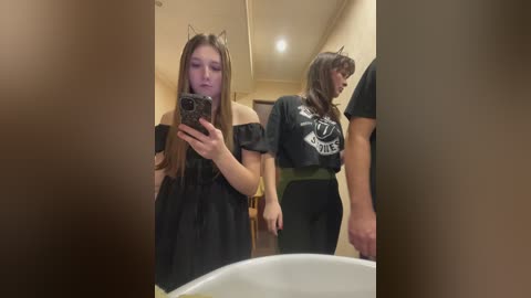 A video of a young woman with long brown hair, wearing black off-shoulder dress and cat ears, taking a selfie in a bathroom mirror. Another person in the background wears a black t-shirt.