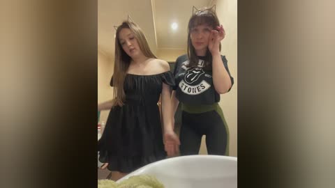 Video of two young women, one in a black off-shoulder dress, the other in a dark green crop top and leggings, both wearing cat ears, in a bathroom.
