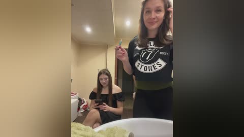 A video captures two women in a bathroom; one, a fair-skinned woman with long brown hair, wears a black T-shirt with a \"U\" logo, and brushes her teeth. The other, a brunette, sits on the counter, reading a book.