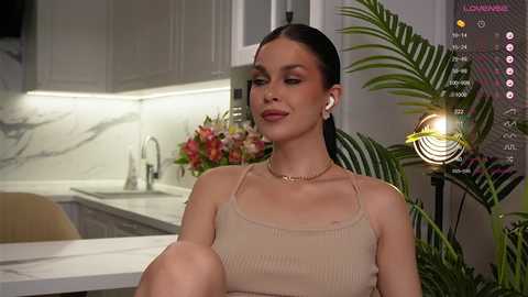 Video of a light-skinned woman with dark hair, wearing a beige ribbed tank top, seated in a modern kitchen with white cabinets, a potted plant, and a digital clock.