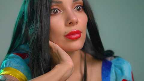 Video of a young woman with long, straight black hair, light skin, and full red lips, wearing a colorful, abstract-patterned blouse, against a muted teal background. She rests her chin on her hand, looking contemplative.