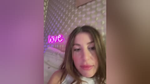 Video of a young Caucasian woman with straight, light brown hair, wearing a white tank top, lying on a bed with a pink heart-shaped neon sign \"love\" and a quilted headboard.