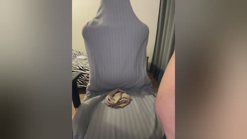 A video of a person sitting on a bed, wearing a grey, ribbed, sleeveless shirt, with a beige bra visible beneath. The background includes zebra-patterned bedding and a dark curtain.