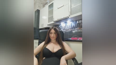 Video of a curvy woman with long brown hair, wearing a black bra, sitting in a modern kitchen with white cabinets and a subway tile backsplash.