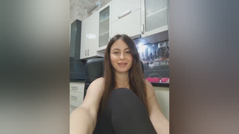 A video of a smiling young woman with long brown hair, wearing a black top, seated in a modern kitchen with white cabinets and black appliances.