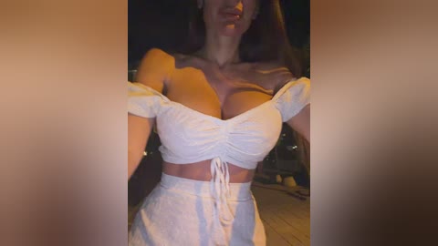 Video of a woman with long, straight brown hair, wearing a white, off-the-shoulder crop top that accentuates her large breasts and high-waisted pants, against a dimly lit, blurry background.