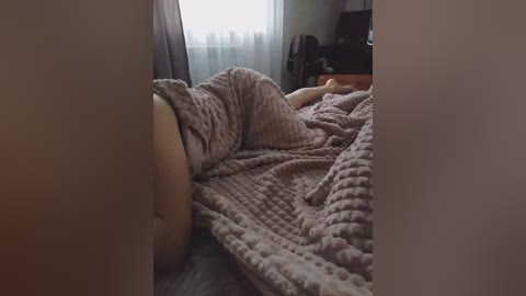 Video of a person lying on a bed, covered in a textured, light pink blanket. The room is dimly lit, with a window showing a cloudy sky outside.