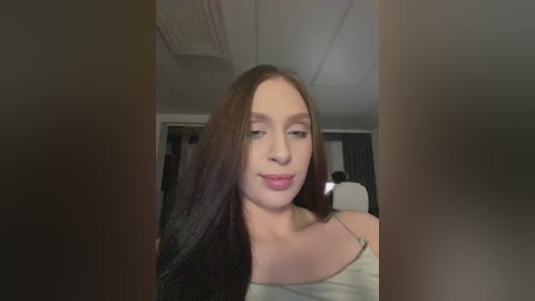 Video of a young Caucasian woman with straight, dark brown hair, light skin, and a slender physique. She wears a light gray, spaghetti-strap top, pouting slightly, in an indoor setting with a white ceiling and dark walls.