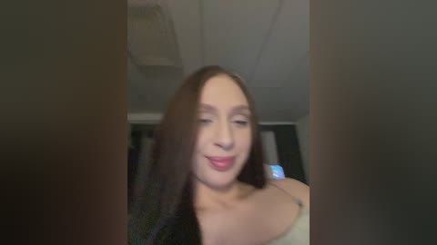 A blurry video of a young woman with straight, dark hair, light skin, and a faint smile. She wears a beige top. The background shows a dimly lit room with a ceiling fan.