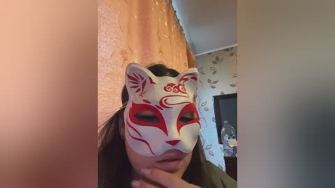 Video of a person wearing a white fox mask with red markings and long black hair, standing in a room with a textured orange wall, patterned wallpaper, and a visible ceiling.