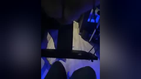 A dimly lit, grainy video showing a close-up of a bed with white sheets and dark objects, possibly a person or a blanket, partially visible. The background is shadowy, with a blueish hue.