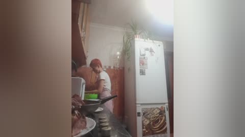 Video of a woman in a kitchen wearing a red headscarf, preparing food on a white stove. Background features wooden cabinetry, a potted plant, and a cluttered countertop.