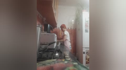 Video of a cozy kitchen with a woman in a red headscarf and white apron cooking on a gas stove. A refrigerator and potted plant are visible in the background.