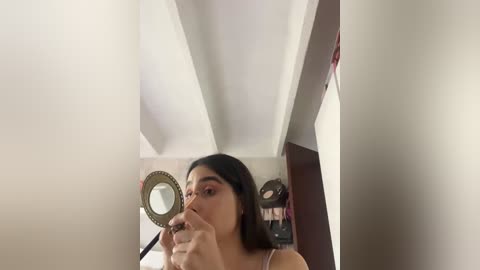 Video of a young woman with light skin, long dark hair, and brown eyes, applying lipstick in a dimly lit room with white walls and framed mirrors.