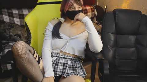 Video of a woman in a white crop top and plaid skirt, seated in a yellow gaming chair, wearing a black mask, with a black-haired woman in fishnet stockings in the background.