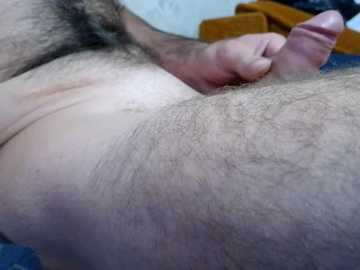 Video of a close-up view of a male's hairy, circumcised penis, being held by a hand, with a blue background and a hint of a brown object in the upper right.