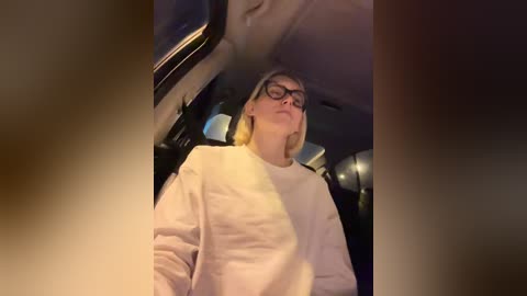 Video of a woman with short, blonde hair, wearing glasses, a white shirt, and a cross-body bag, seated in a car at night with dim, ambient lighting.