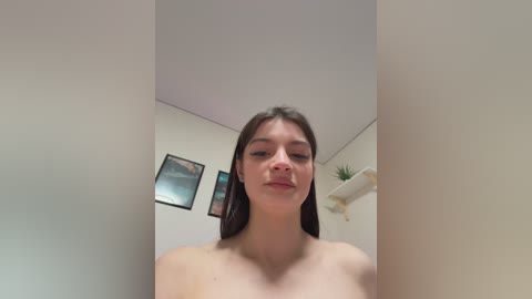 Video of a young Caucasian woman with straight brown hair, standing topless indoors. The background features minimalist decor with framed pictures and a shelf.