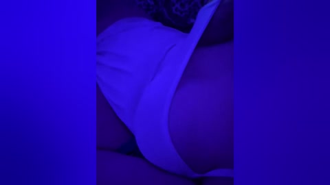 A close-up video under dim blue lighting shows a woman's bare chest and cleavage, partially covered by a white bra strap. The image is grainy and slightly blurred, with shadows emphasizing the intimate, sensual atmosphere.