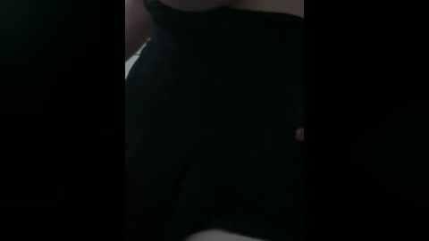A close-up, dimly lit video of a person's torso in black clothing, with a dark background. The focus is on the waist and upper thighs, emphasizing the contours of the figure.