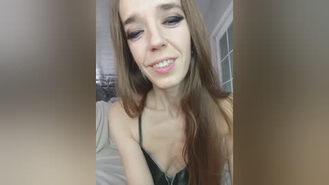 Video of a young woman with fair skin, long brown hair, and blue eyes, wearing a black lace bra, smiling slightly, in a softly lit bedroom.