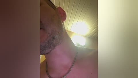 A close-up video of a bearded man with a silver necklace, taken from a low angle, showing his profile and the ceiling with a light fixture.