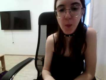 Video of a young, fair-skinned woman with glasses, black hair, and a black choker, wearing a black top, sitting in a black office chair in a modern, minimalist room with a large flat-screen TV on the wall.