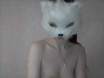 Video of a topless person with a white fox mask, wearing a dainty necklace, against a plain, grayish background. The individual's face is partially obscured by the mask, creating a mysterious and surreal atmosphere.