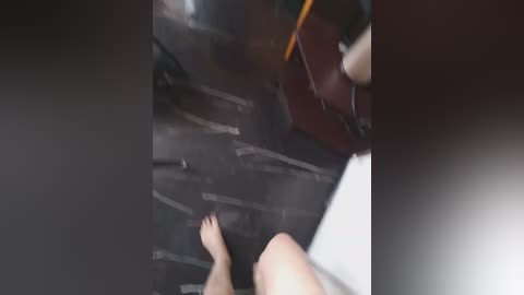 A blurry video showing a barefoot person walking on a dark, reflective floor with vertical white lines. The person's legs are visible, and a blurred figure in the background carries a guitar case.