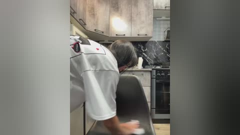 Video of a man with short gray hair, wearing a white chef's jacket, bent over a black kitchen island, preparing food in a modern, stainless steel kitchen with granite countertops and wooden flooring.