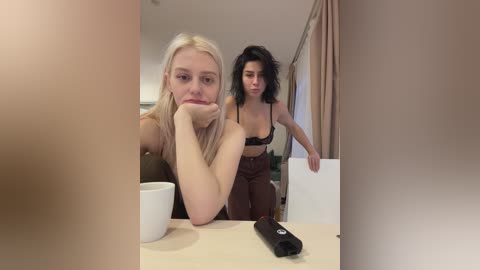 Video of a blonde woman with a pensive expression, sitting at a table in a beige room, with a black remote and a white cup in front of her.