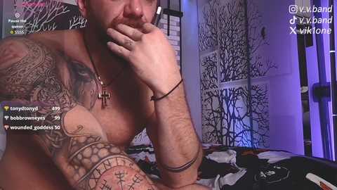 Video of a shirtless, tattooed man with a beard, leaning on a bed with a floral-patterned blanket, wearing a cross necklace, in a dimly lit room with a mural of trees.