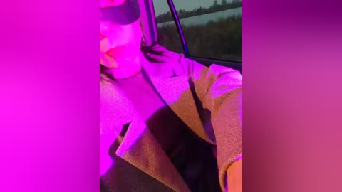 Video of a person wearing a black blindfold, seated in a car, with purple light illuminating their face and shoulders, viewed through a window overlooking a dark, wooded area.