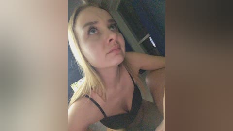 Video of a young blonde woman with light skin, wearing a black bra, looking up with a puzzled expression. Background includes a dark, blurry room with a window and a computer monitor.