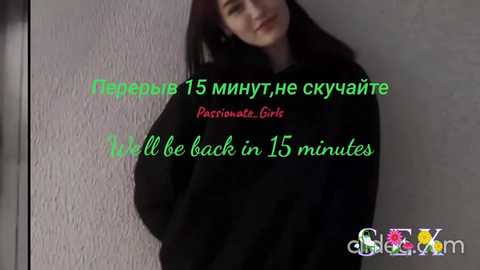 Video of a young woman with long black hair, wearing a black coat, smiling slightly. Text overlays in green and red: \"\u0422\u0435\u043f\u0435\u0440\u044c 15 \u043c\u0438\u043d\u0443\u0442, \u043f\u043e\u043a\u0430 \u0434\u0435\u0432\u043e\u0447\u043a\u0430 \u0432 15 \u043c\u0438\u043d\u0443\u0442.\