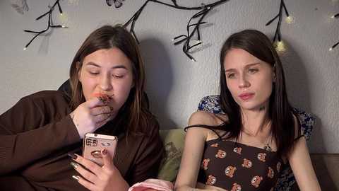 Video of two young women with straight, dark hair, wearing dark sweaters, eating ice cream and looking at their phones, in a cozy indoor setting with string lights and floral pillows.