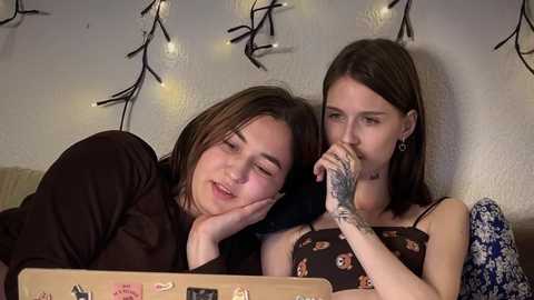 Video of two young women with short brown hair, lying on a sofa, one with a tattooed arm, wearing a floral dress, against a wall with string lights and a floral pillow.