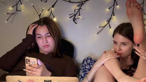 Video of two young women in casual attire, one on a laptop, the other playfully biting her foot, against a festive string-lit wall.