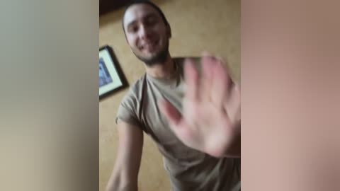 Video of a smiling, light-skinned man with short hair in a beige shirt, slightly blurred, standing in a room with a framed picture on a light brown wall.