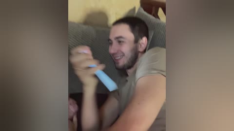 A video captures a smiling man with short dark hair, holding a blue controller, sitting on a grey couch, with a blurred figure's legs in the foreground.