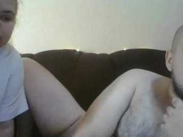 Video of a bearded man with a tattooed arm, sitting shirtless on a couch, and a woman with dark hair partially visible, both looking at a TV screen.