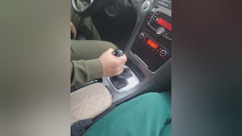 A video of a person's hand turning a car's gear stick, wearing a green jacket, beige pants, and a green blanket covering their legs. The car's interior shows a dark dashboard with a red display and steering wheel.
