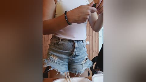Video of a woman in a white tank top and distressed denim shorts, holding a phone, with a black beaded bracelet on her wrist. Background features wooden walls and a white door.