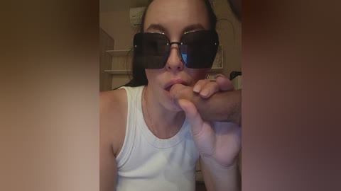 Video of a young woman with light skin, dark hair, and large black sunglasses, kneeling, licking a man's erect penis in a dimly lit room with beige walls and a white shelf in the background.