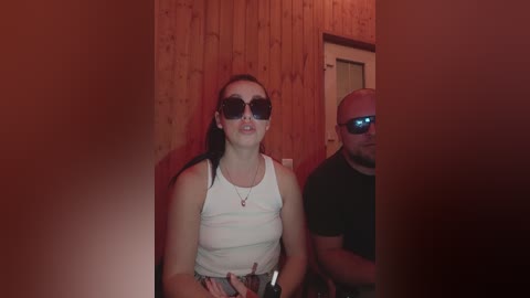 Video of a fair-skinned woman with long dark hair, wearing sunglasses and a white tank top, standing next to a bald man in a black t-shirt in a dimly-lit wooden room.
