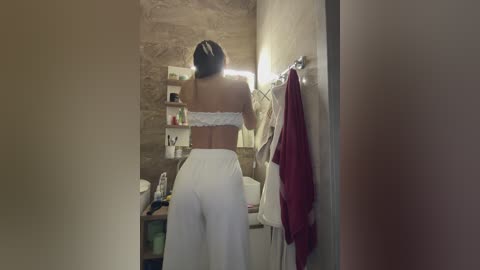 Video of a woman in white lace bra and high-waisted pants, standing with back to camera, in a rustic bathroom with stone walls and shelves, red towel hanging on a hook.