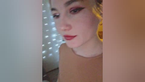 Video of a young woman with fair skin, red lipstick, and bold winged eyeliner, wearing a beige top, in a dimly lit room with string lights and yellow headphones.